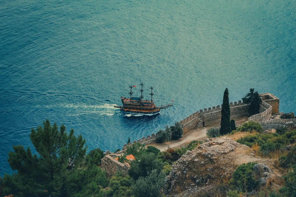 Aerial shot of a pirate ship sailing near Alanya Castle's scenic coastal cliff.
